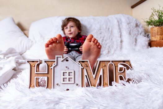 Happy Kid boy plays games with controller and lying on the bed at home and relax. Stay at home safety quarantine coronavirus COVID-19 pandemic prevention. Wooden house. Barefeet kid on the bed