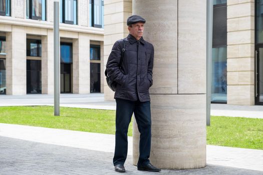 Adult Caucasian male stands near modern building with columns. Pensioner in seasonal clothes while walking around the city. Selective focus.