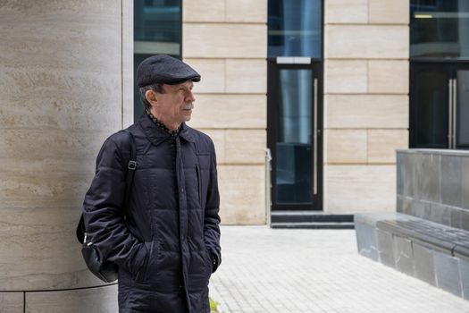 Adult Caucasian male stands near modern building with columns. Pensioner in seasonal clothes while walking around the city. Selective focus.