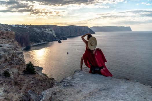 A girl with loose hair in a red dress and hat stands on the rocks above the sea. In the background, the sea and the rocks. The concept of travel