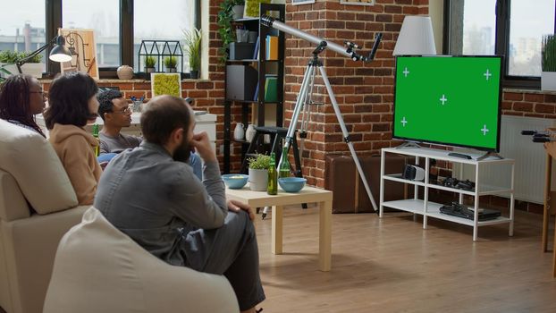 Men and women watching television with green screen in living room, using isolated chroma key template with mockup copy space and blank background. Modern technology on TV display. Tripod shot.