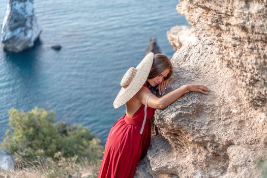 A girl with loose hair in a red dress and hat stands on the rocks above the sea. In the background, the sea and the rocks. The concept of travel