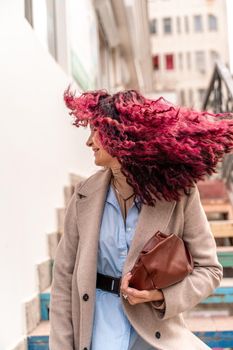 A woman with pink curly hair blowing in the wind. Standing on the stairs in a light raincoat and blue shirt, bag in hand