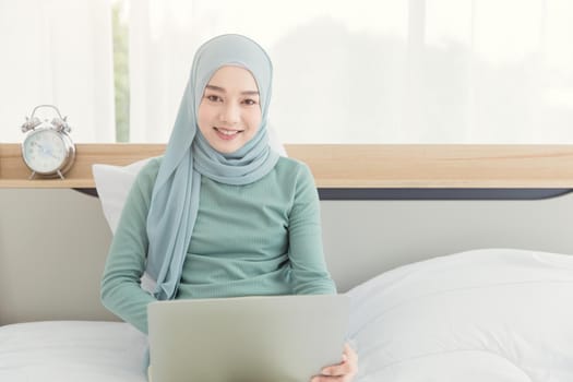 Asian Muslim Islamic woman with hijab happy enjoy online remote work at home with her laptop on the bed.
