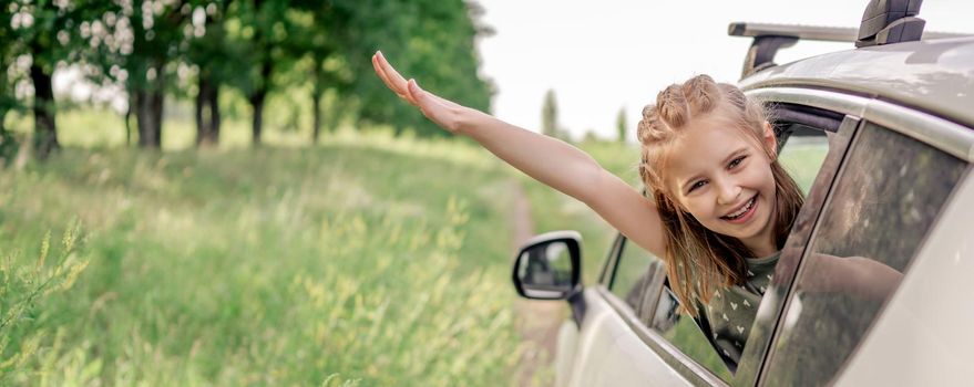 Adorable preteen girl sitting in the car and looking out the window open and smiling. Portrait of happy child kid in the vehicle outdoors in the field during summer trip