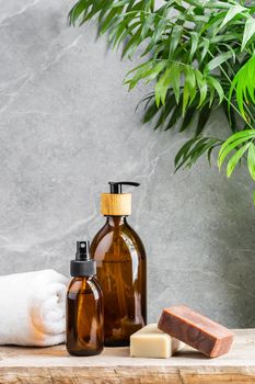 Bathroom table with white towel, dark glass bottles with liquid skin care products and natural handmade solid soap bars over grey marble background and green plant above