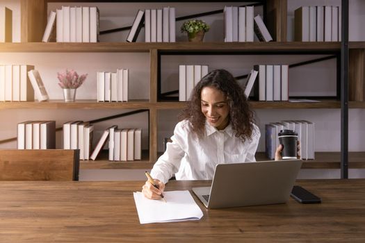 Author working women creative enjoy happy writing new idea on paper. African American black female sitting work journalist at wooden table with book shelf background.