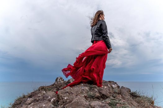 A woman in a red dress and leather jacket stands above a stormy sky, her dress fluttering, the fabric flying in the wind