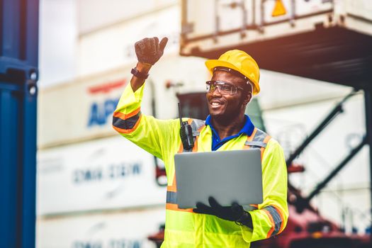 Black African happy worker working in logistic shipping with laptop computer control loading containers at port cargo. Import export shipping industry people.