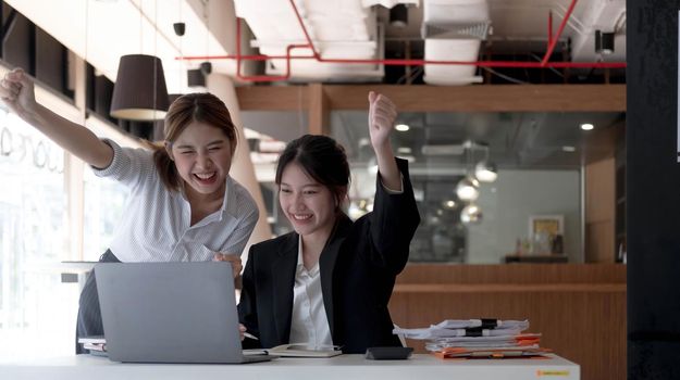 Two young Asian businesswomen show joyful expression of success at work smiling happily with a laptop computer in a modern office..