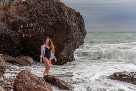 A beautiful girl in a black dress is walking on the waves, big waves with white foam. A cloudy stormy day at sea, with clouds and big waves hitting the rocks