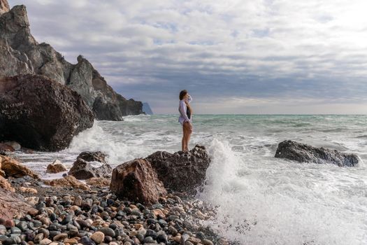 A beautiful girl in a white shirt and black swimsuit stands on a rock, big waves with white foam. A cloudy stormy day at sea, with clouds and big waves hitting the rocks