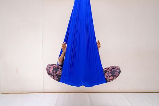 A young woman poses while doing anti-gravity aerial yoga in a blue hammock on a white background