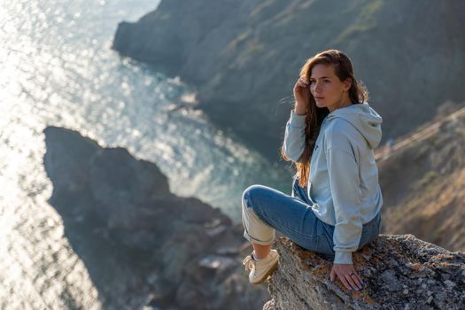 Woman tourist enjoying the sunset over the sea mountain landscape. Sits outdoors on a rock above the sea. She is wearing jeans and a blue hoodie. Healthy lifestyle, harmony and meditation.