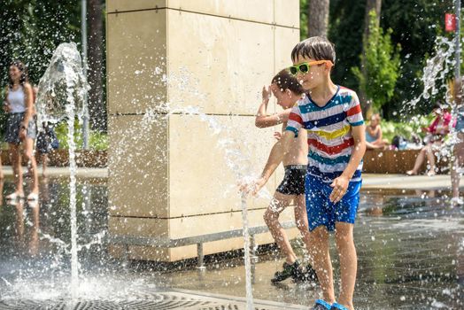 Kyiv, Ukraine - August 01, 2021: Boys jumping in water fountains. Children playing with a city fountain on hot summer day. Happy friends having fun in fountain. Summer weather. Friendship, lifestyle and vacation.