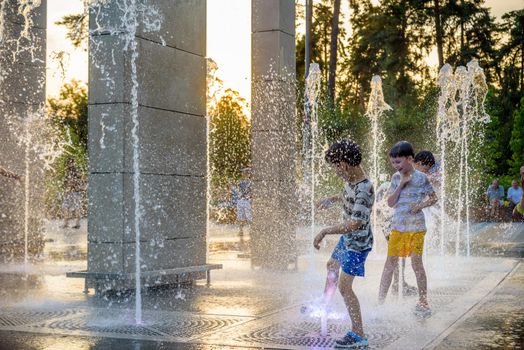 Kyiv, Ukraine - August 01, 2021: Boys jumping in water fountains. Children playing with a city fountain on hot summer day. Happy friends having fun in fountain. Summer weather. Friendship, lifestyle and vacation.