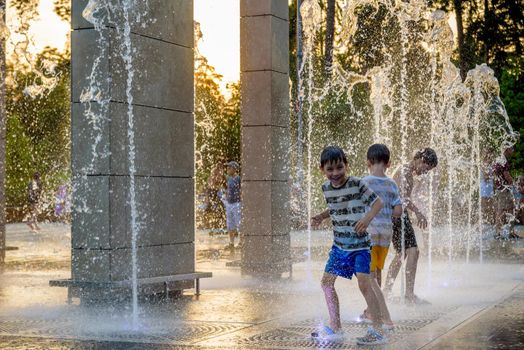 Kyiv, Ukraine - August 01, 2021: Boys jumping in water fountains. Children playing with a city fountain on hot summer day. Happy friends having fun in fountain. Summer weather. Friendship, lifestyle and vacation.