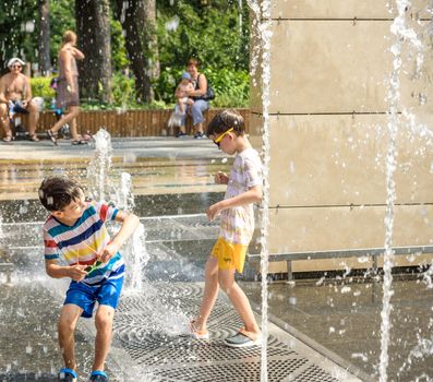 Kyiv, Ukraine - August 01, 2021: Boys jumping in water fountains. Children playing with a city fountain on hot summer day. Happy friends having fun in fountain. Summer weather. Friendship, lifestyle and vacation.