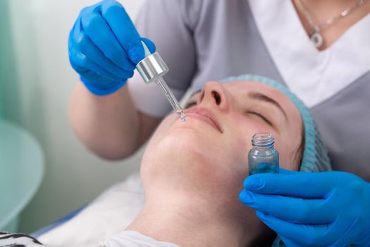 Hands of cosmetologist dripping nourishing oil facial skin of young woman in spa salon. Facial treatment and skincare concept.