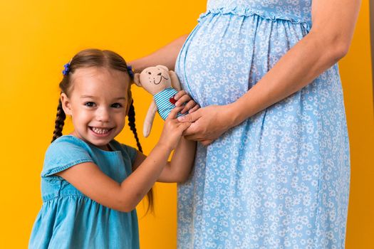 Motherhood, love, Childhood, pregnancy, hot summer - croped portrait pregnant unrecognizable mother woman blue dress little preschool Daughter girl sibling teddy bear hug mum belly on yellow background.