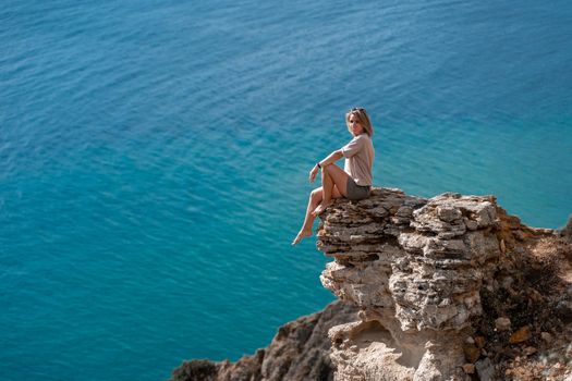 The summer lifestyle of a happy stunning woman sitting on a rock above the sea. Enjoying life and looking at the sea. Turquoise sea background. In a red bathing suit and a white shirt