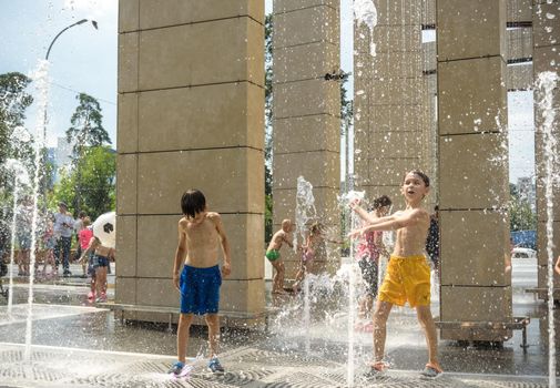 Kyiv, Ukraine - August 01, 2021: Boys jumping in water fountains. Children playing with a city fountain on hot summer day. Happy friends having fun in fountain. Summer weather. Friendship, lifestyle and vacation.