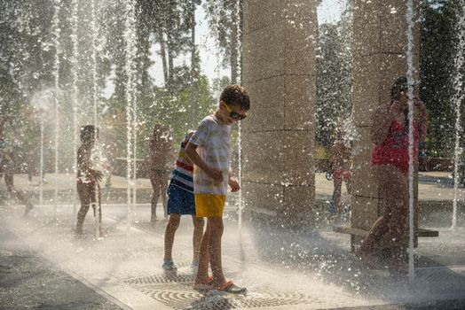 Kyiv, Ukraine - August 01, 2021: Boys jumping in water fountains. Children playing with a city fountain on hot summer day. Happy friends having fun in fountain. Summer weather. Friendship, lifestyle and vacation.