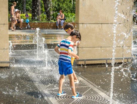 Kyiv, Ukraine - August 01, 2021: Boys jumping in water fountains. Children playing with a city fountain on hot summer day. Happy friends having fun in fountain. Summer weather. Friendship, lifestyle and vacation.