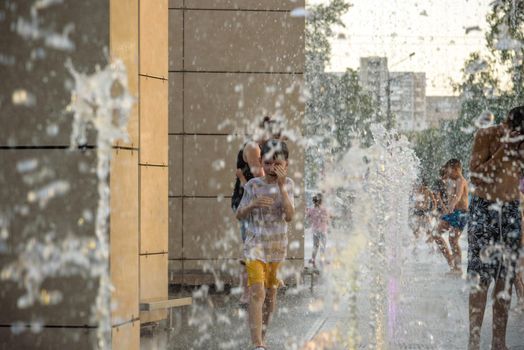 Kyiv, Ukraine - August 01, 2021: Boys jumping in water fountains. Children playing with a city fountain on hot summer day. Happy friends having fun in fountain. Summer weather. Friendship, lifestyle and vacation.