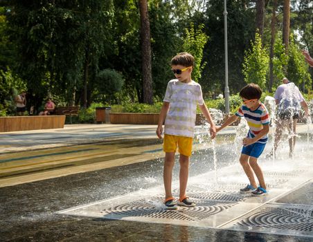 Kyiv, Ukraine - August 01, 2021: Boys jumping in water fountains. Children playing with a city fountain on hot summer day. Happy friends having fun in fountain. Summer weather. Friendship, lifestyle and vacation.