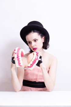 A woman in a dress and hat sits at a table and holds a mock-up of her teeth. High quality photo