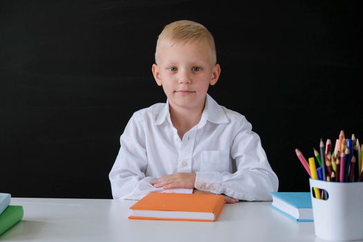 Cute little boy sitting at the table on blackboard backgrpund. Child from elementary school. Education concept.