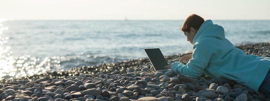 Caucasian woman lying on her stomach on a pebble beach and typing on a laptop. Remote work freelancer