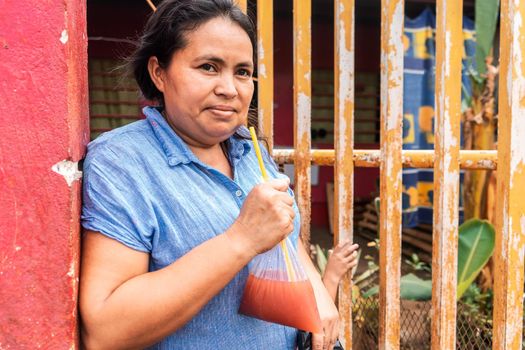 Latin woman from Nicaragua drinking a fruit drink in a plastic bag typical of Latin America. He is outside his house and looking at the camera.