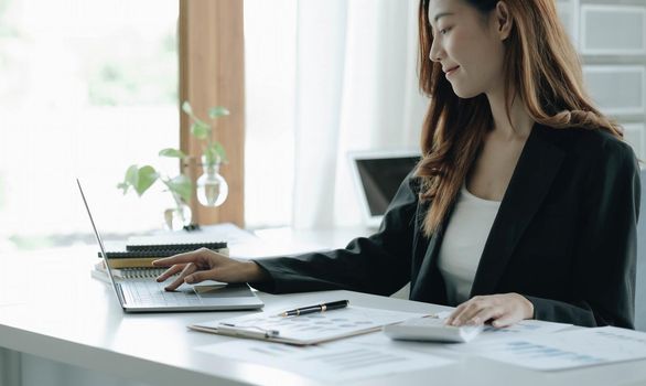 Beautiful Asian woman sitting in office using laptop and calculator. Happy business woman sitting at a desk in an office with a tablet computer.