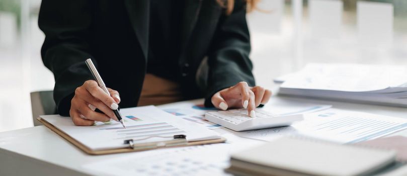 Close up of businesswoman or accountant hand holding pen working on calculator to calculate business data, accountancy document and laptop computer at office, business concept.