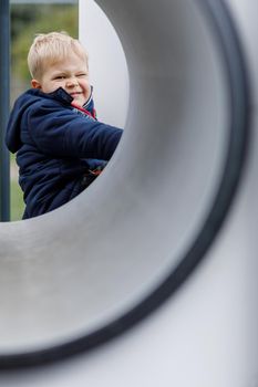 The little boy poses at the end of the tunnel with one eye closed. Fun games on the playground, carefree childhood