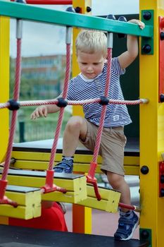 Little boy with striped shirts goes by the monkey bridge at public  playground. Brave child. Risky steps. Use handrails