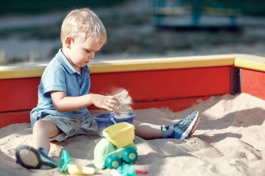 Toddler boy is playing in a red and yellow edge sandbox with sand toys. Busy child, wearing blue shirt is dropping sand into a blue bucket. Childhood joys outdoor