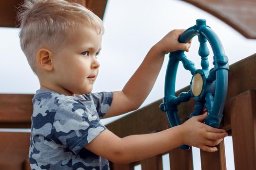 Cheeky child is driving a ship wheel in a playground. Kid wearing camouflage feels cool in a wooden playhouse. Childhood full of adventure