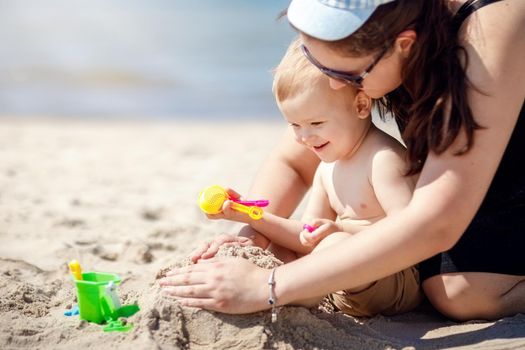 The mother plays with her son by the sea, she teaches the child to build a sand castle. Mother's care, and love, a happy child smiles. Travel with young children. Family summer vacation.
