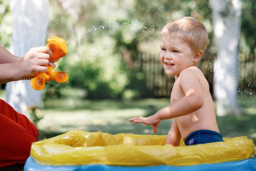 Beautiful boy plays in a garden pool at hot summer day. Splashing with water. Trying to avoid water. Childhood happiness in a sunshine