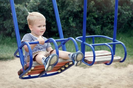 Little boy swinging on the swings of a public playground. Looking at somewhere and dreaming. Happy childhood in outdoor