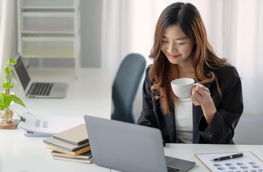 Smiling Asian businesswoman holding a coffee mug and laptop at the office.