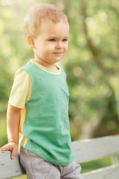 Well-meaning, happy boy rests in the garden, he calmly watches the environment and smiles. Sunny summer day, cheerful child with green shirt, yellow sleeves.