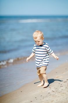 Barefoot little boy walks on the seaside at noon near the water. Child on vacation in summer at the sea on good weather.