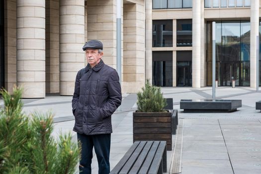 Adult Caucasian man stands near modern building with columns. Pensioner in seasonal clothes during a walk in the city. Selective focus.