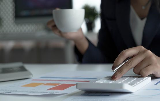 Asian businesswoman working on laptop computer and drinking coffee in modern office near window with calculator and business report on desk. work from home concept