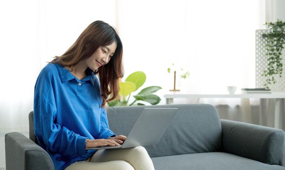 Young business freelance Asian woman working on laptop checking social media while lying on the sofa when relax in living room at home.