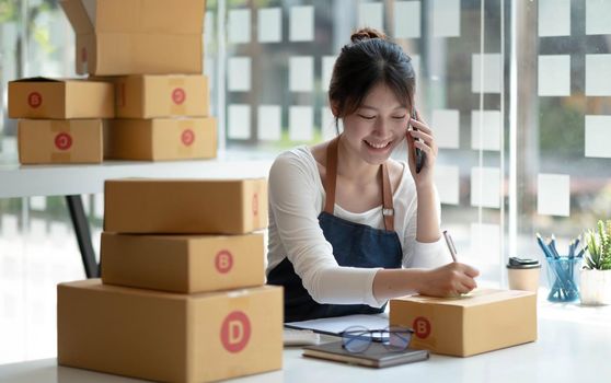 Happy young Asian business owner woman prepare parcel box and check online orders of product for deliver to customer on smartphone. Shopping Online concept.